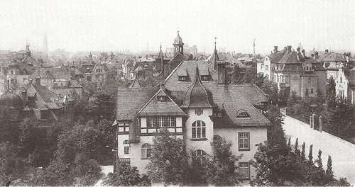 Die Möhlstraße vom Firsthäuschen der Villa Wannieck (HsNr. 32) aus nach Süden. Im Vordergrund die große Doppelvilla der Rentiere Klara Herz (HsNr. 30 und 30a). Rechts noch im Bild die Villa von Julius Kaufmann (HsNr. 21) und der mächtige Stock der Villa des Fabrikdirektors Hans Kullen (Hs.Nr. 19). Schemenhaft im Hintergrund links die Pfarrkirche St. Johann Baptist von Haidhausen, das Maximilianeum (Mitte) und rechts der Friedensengel. Quelle: Buchscan aus: s.o.